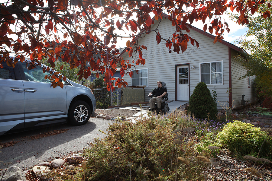 A green-shirted man travels in a power wheelchair down the wooden ramp toward his van. Red leaves of a tree are visible in the foreground, and green and purple shrubs border the lawn and driveway.